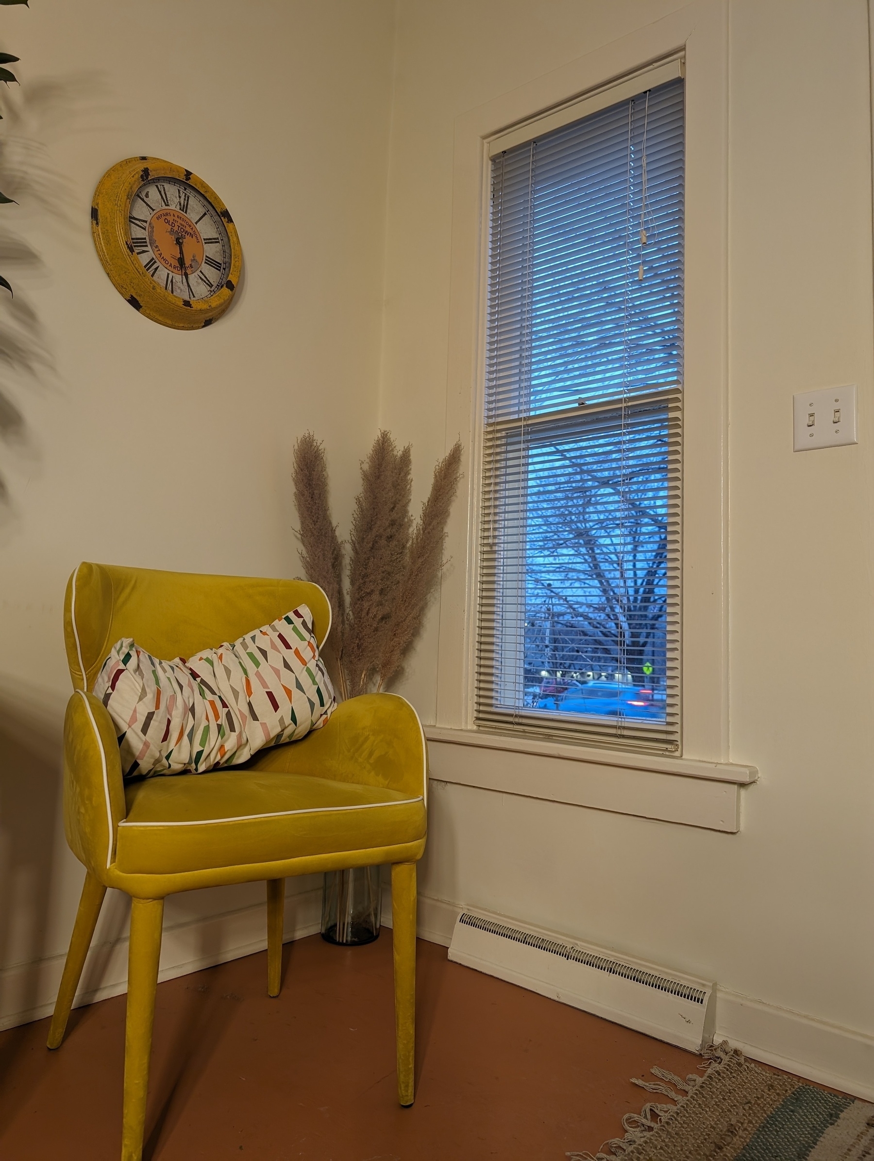 A yellow chair with low and small wraparound wings sits on a rust colored floor. It is in the corner of a room, next to a window with shades that are opened. Above the chair is an analog clock, the time is approximately 5:25 PM.