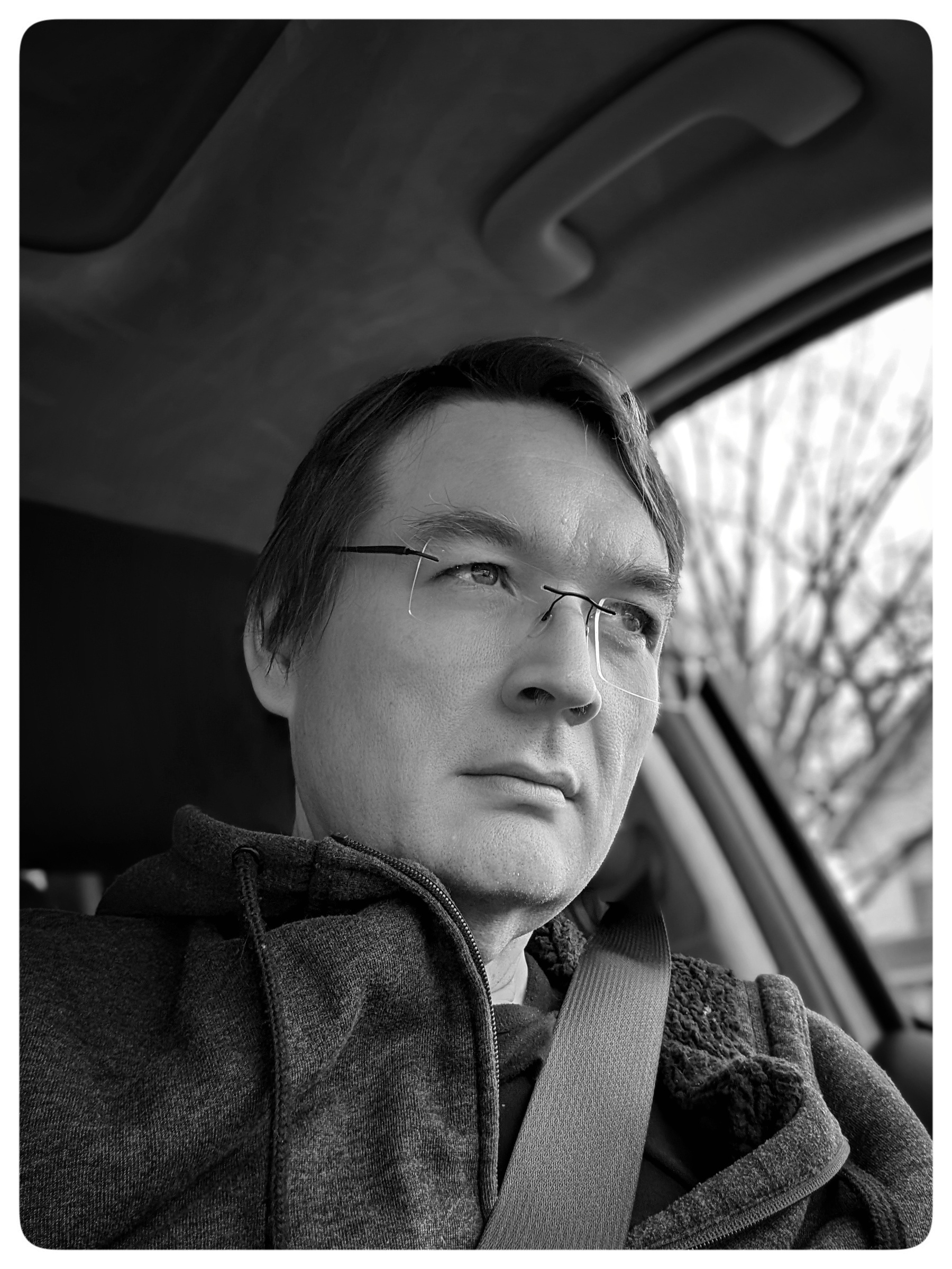 A black and white selfie image of myself. I'm sitting in my vehicle, not looking at the camera but instead looking forward, out of the front window. My seatbelt is fastened.