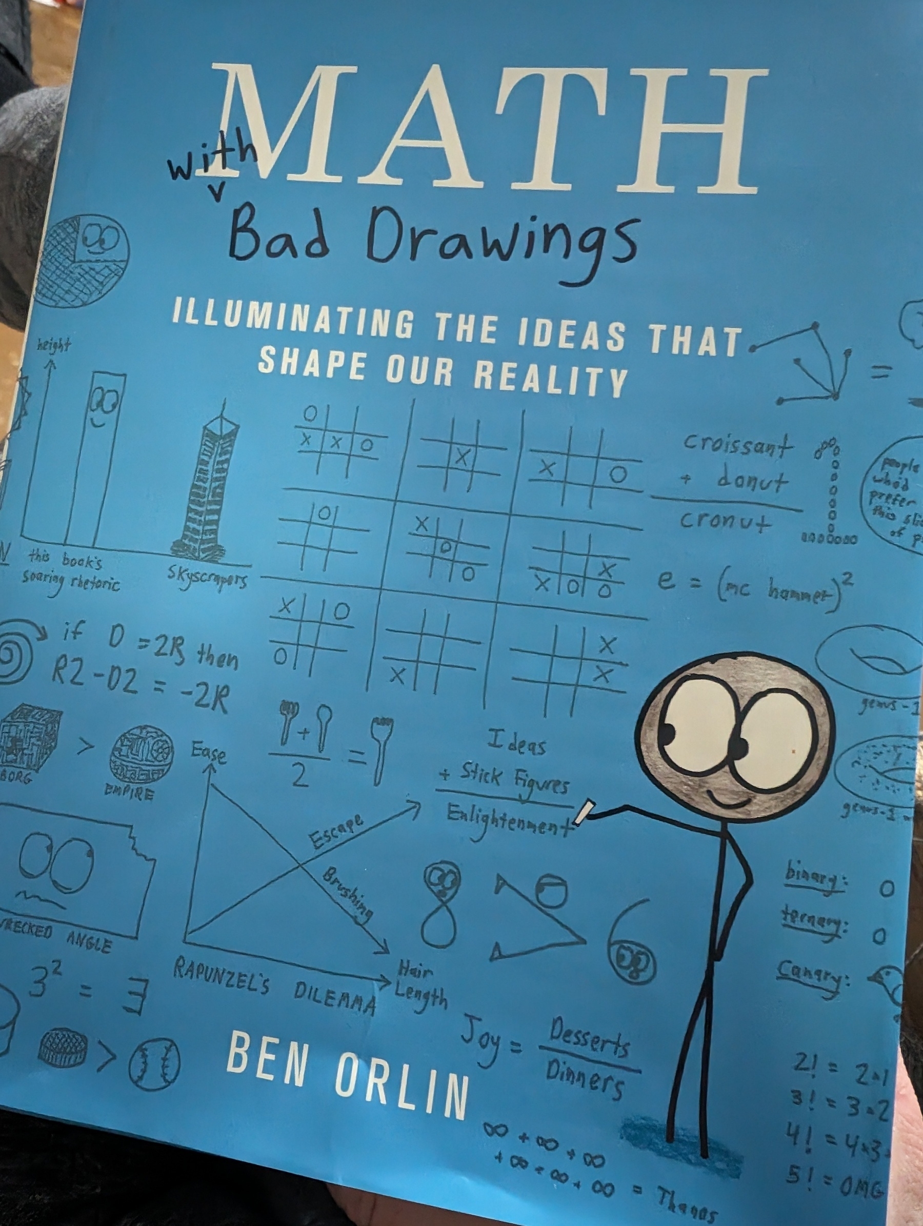 Photo of "Math with Bad Drawings: Illuminating the ideas that shape our reality" by Ben Orlin. The cover is filled with whimsical hand drawn (the author of the book would describe it as badly drawn I'm sure) artwork of various items (tic tac toe, graphs, equations, etc). A stick figure holding a piece of chalk is prominently featured.