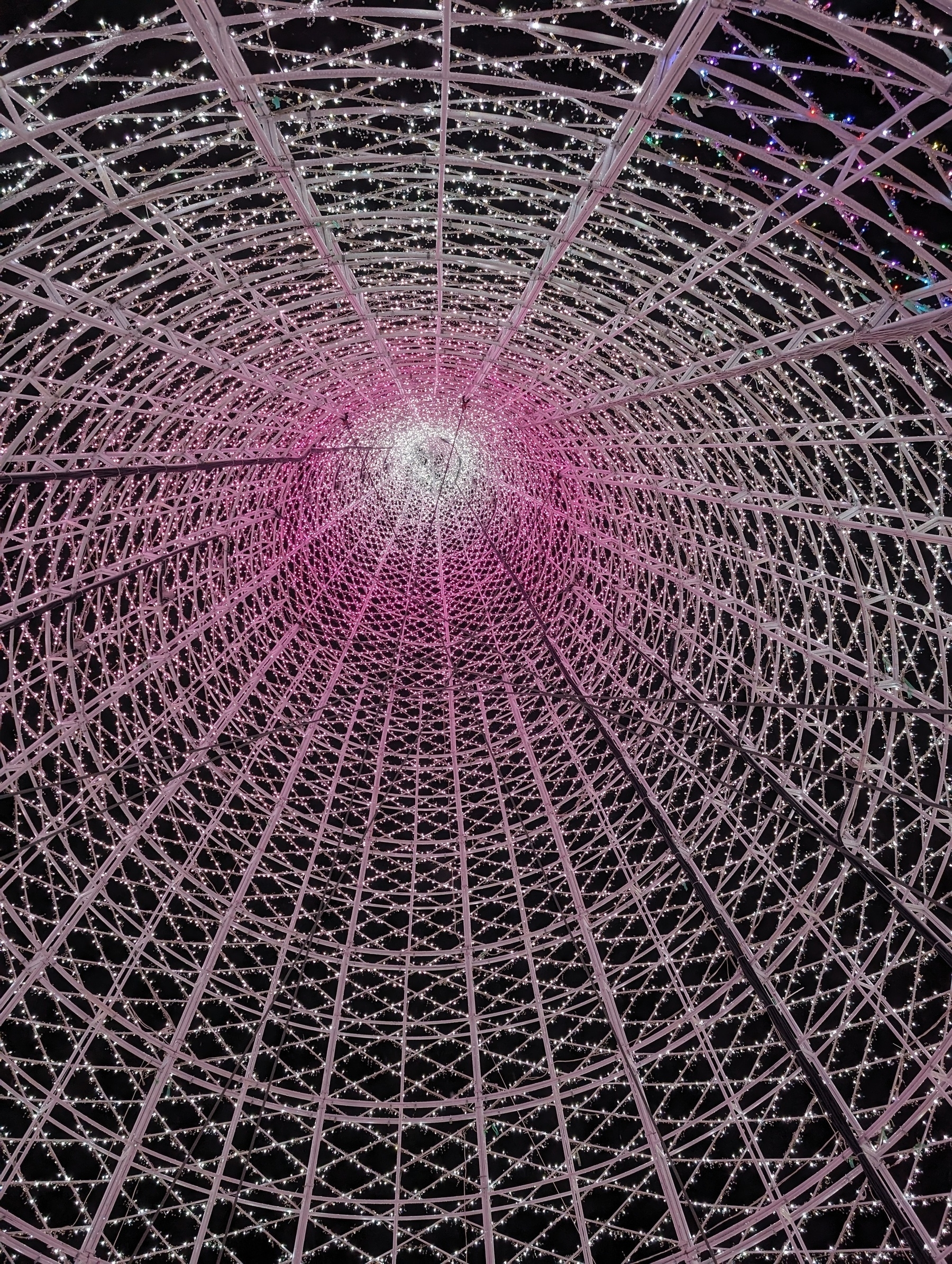 A photo taken from within the interior of a wireframe holiday tree, looking up to the apex of the tree structure. It is illuminated and towards the center of the image is a bright white/violet burst of light.