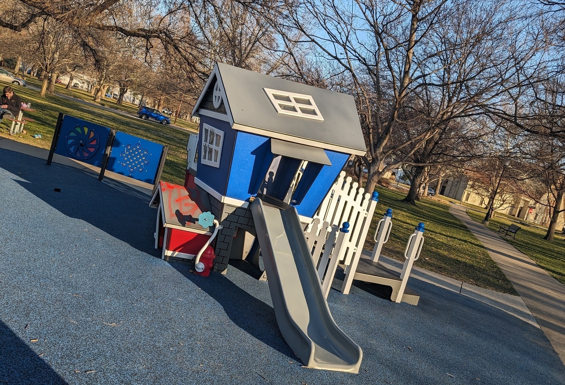 Photo of a children's playhouse at a park. The walls of the home are all at interesting angles. There is a small slide exiting the near wall.