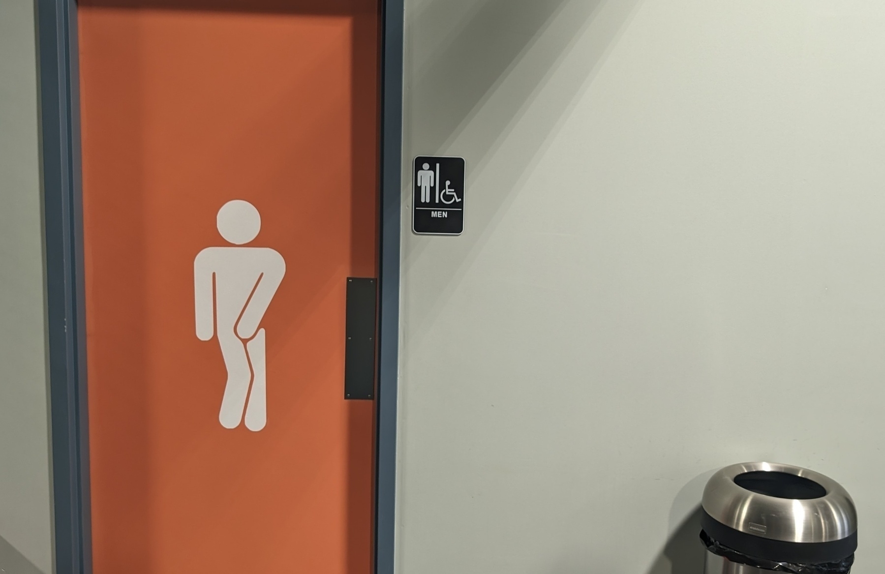 Photo of a restroom door. The door is orange in color, a stark contrast to the paler white color of the wall. Featured on the door is a figure that indicates what could be referred to as a youngster doing the "gotta potty" dance.