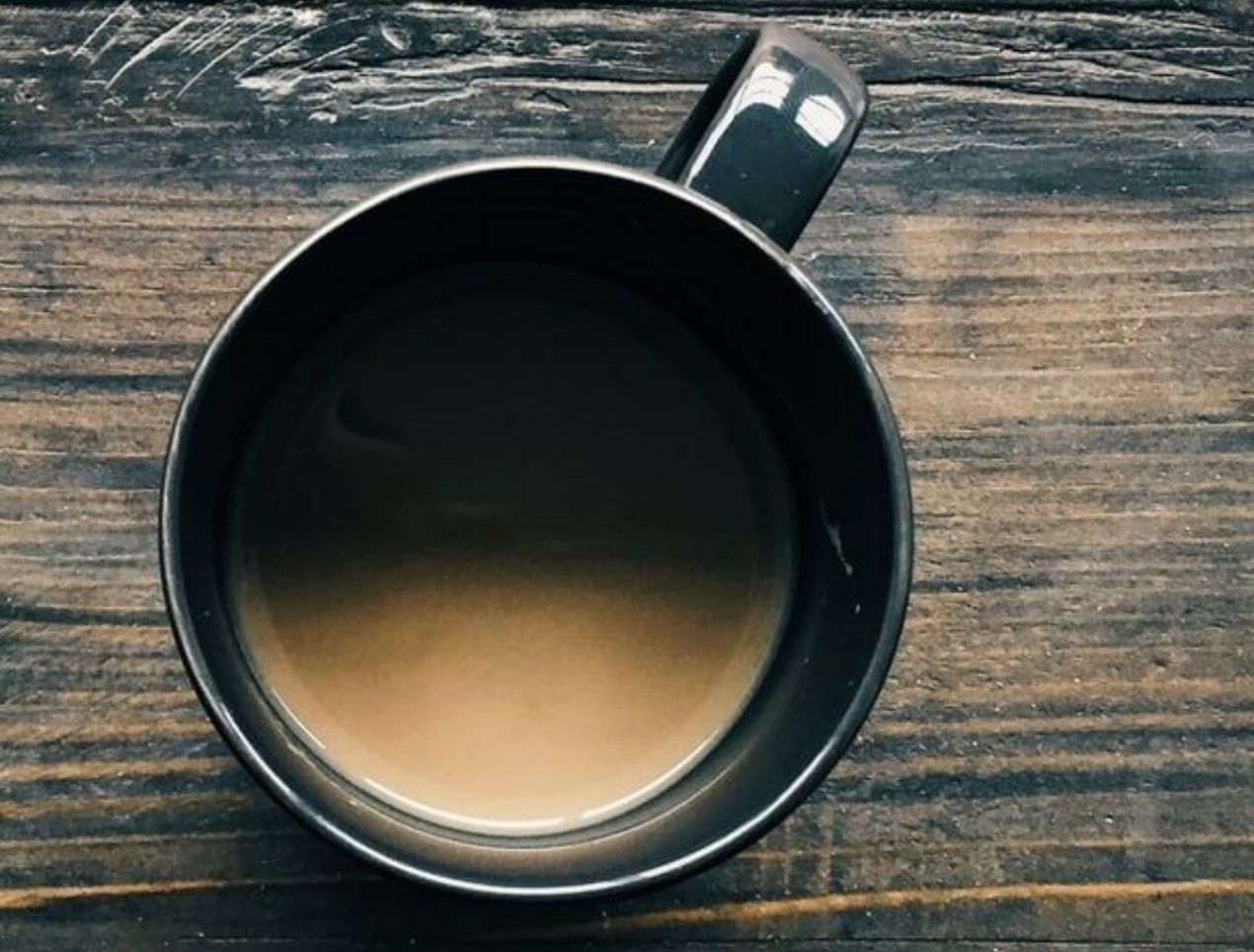 Image of what I believe to be a cup of coffee with cream, sitting on a dark wooden table. An overhead shot.