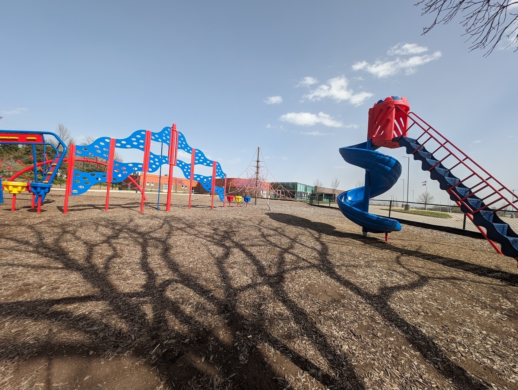 Photo of a local school playground. Red and blue jungle gym climbing equipment. The foreground features a windy tree shadow.