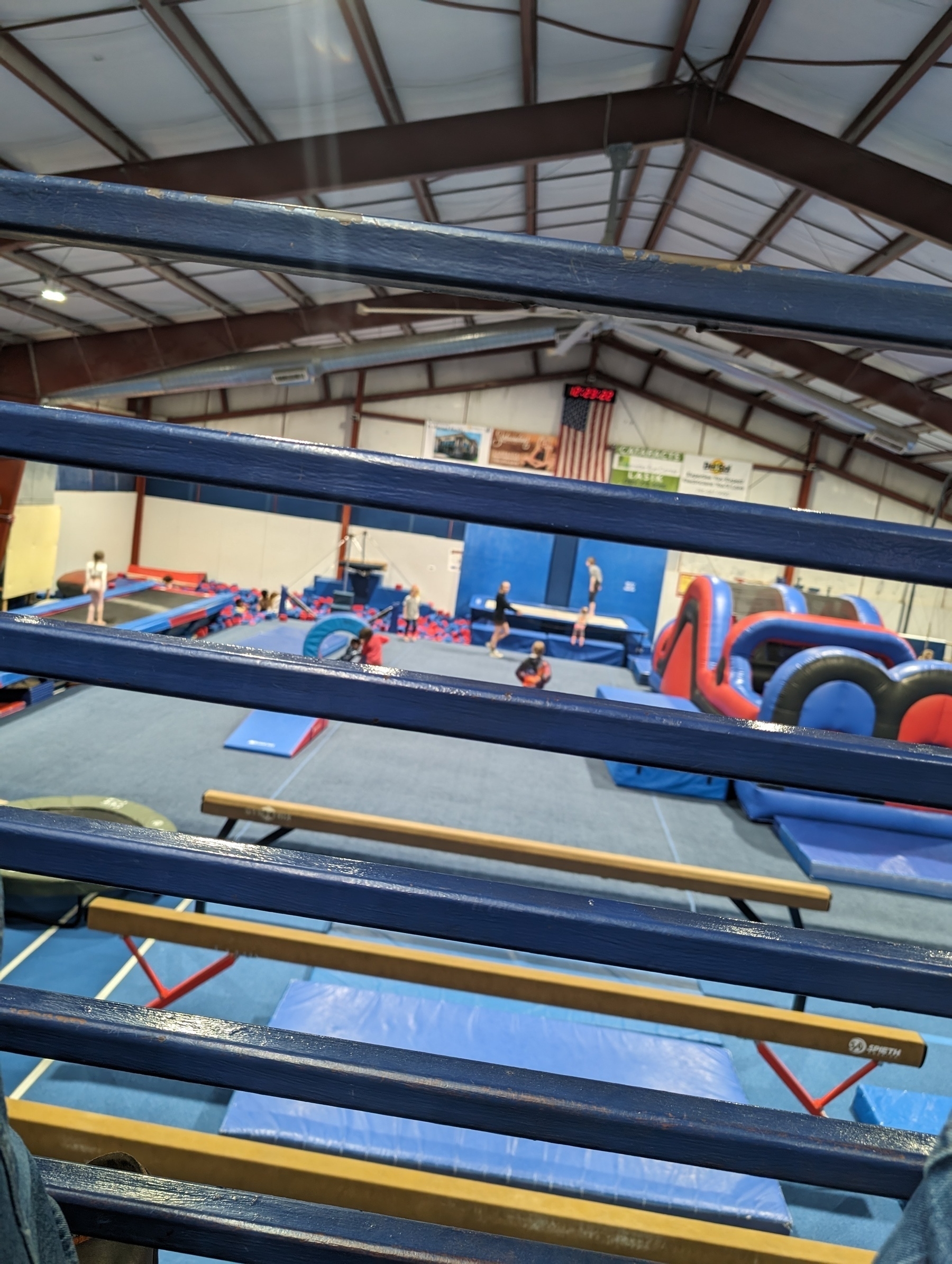 A view from the upper level of a local gymnastics center, overlooking children at play during a birthday party. There is a long trampoline to the left, ending in a foam block pit. There are ramps and bars in the central area. At the far end is another trampoline and bar, to the right of the foam pit. The main feature on the right side is a bounce house. Situated immediately in front of the viewer are the metal railings to keep one from falling.