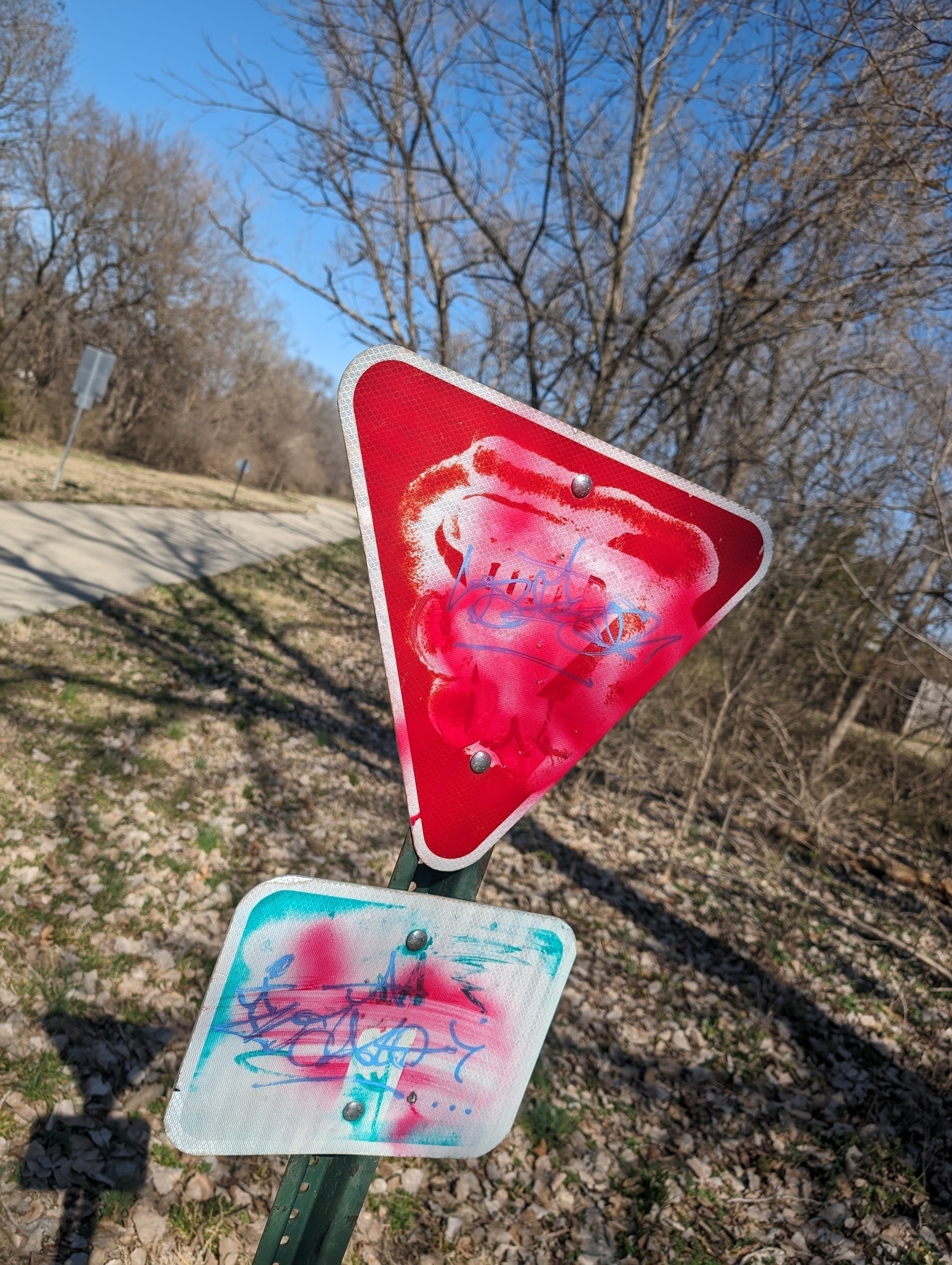 Photo of a Yield sign that has been spray painted over with more red. Beneath that is another unknown sign that is also spray painted over.
